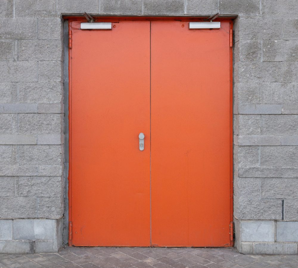 Gray brick wall with wide bright orange door in the centre of the photo. Entry into building.